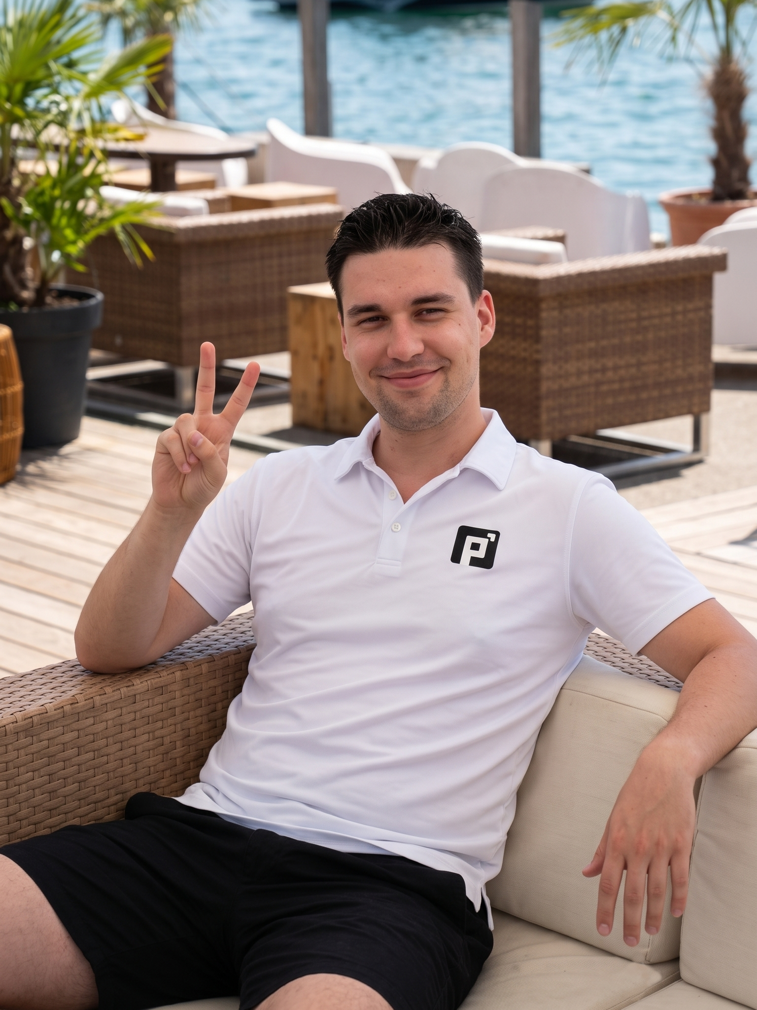 A man sitting on a beige outdoor patio sofa wearing a white short-sleeve polo shirt with a black FootJoy logo on the left chest, paired with black shorts. The background shows a wooden deck, wicker furniture, and a blue ocean view.
